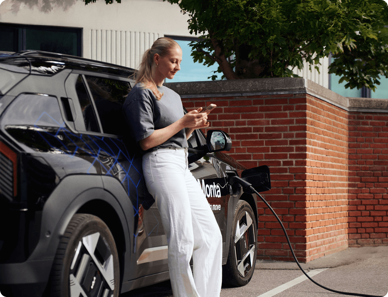 A woman stands beside an electric car, looking at her phone while it charges.