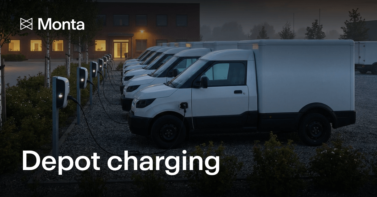 A row of white electric vans charging at a depot, with charging stations in a gravel area and a building in the background.