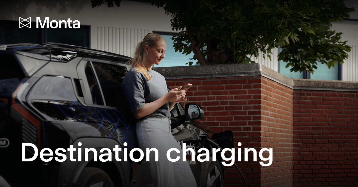 A woman stands beside a black electric vehicle, checking her phone, with a brick wall and trees in the background. Text reads "Destination charging".