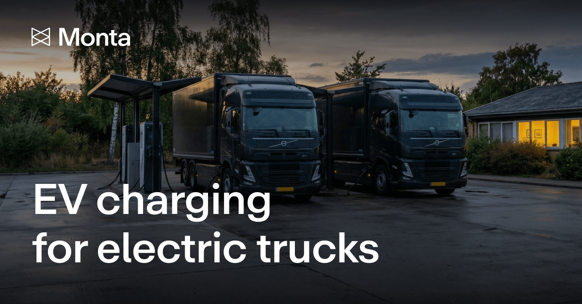 Two electric trucks parked at a charging station during dusk, with a well-lit building in the background and trees surrounding the area.