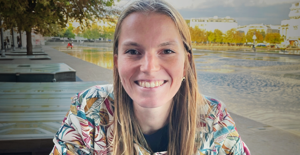 A smiling woman with long blonde hair wearing a floral jacket, seated outdoors near a waterfront plaza