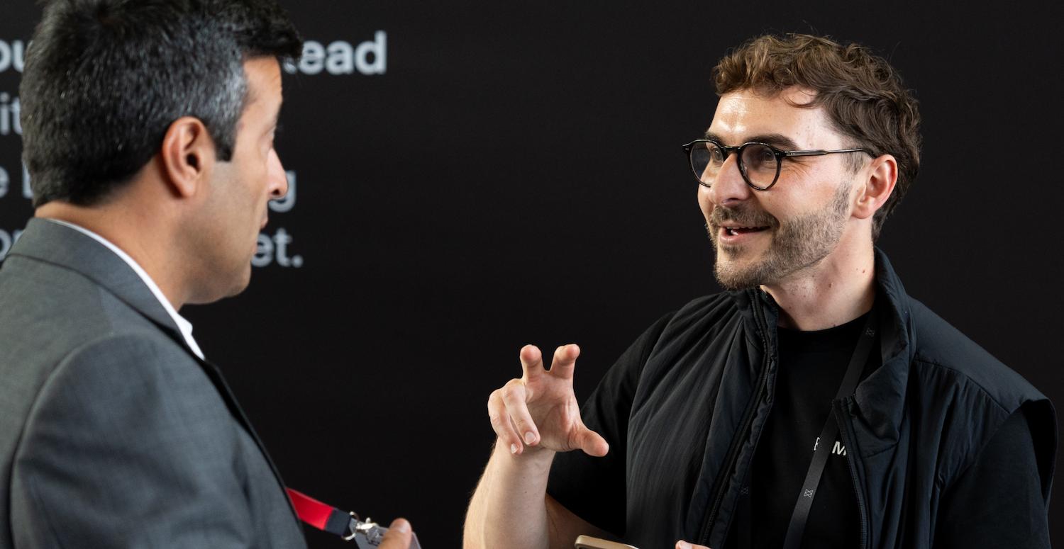 Two men engaged in conversation. One in a grey suit, the other in a black vest with glasses, gesturing animatedly against a dark background.