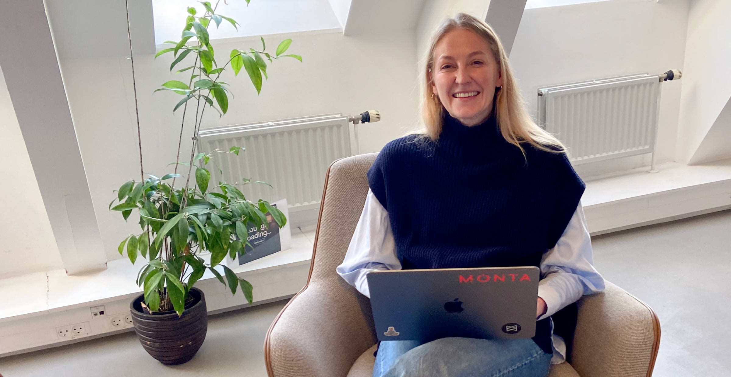 Smiling woman with long blonde hair sits in a comfy chair, wearing a black turtleneck and blue jeans, laptop on her lap, indoor plant beside her.