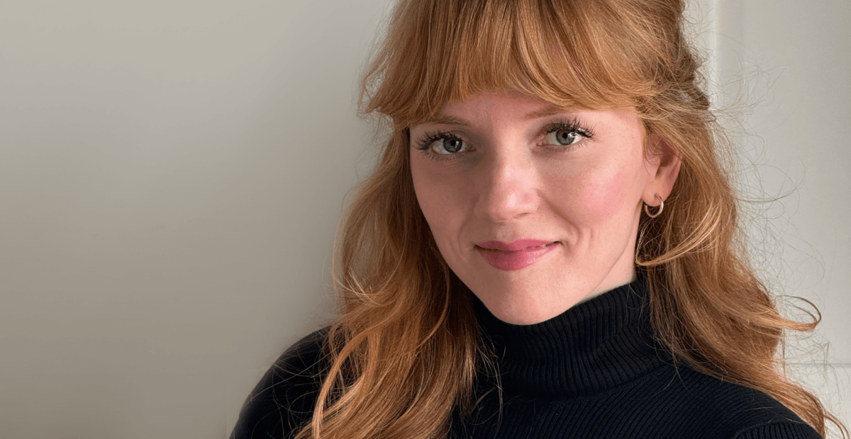 Portrait of a woman with long, wavy red hair, wearing a black turtleneck, smiling softly against a neutral background.