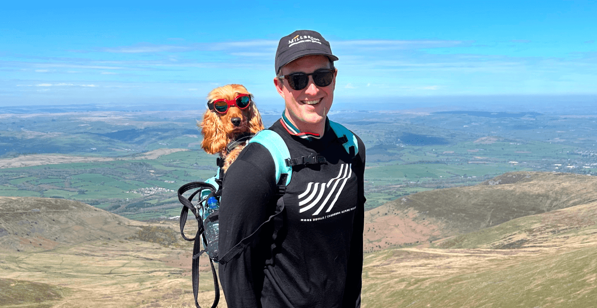 Man wearing sunglasses and a backpack stands on a mountain, with a dog in sunglasses nestled in the backpack, against a scenic landscape.
