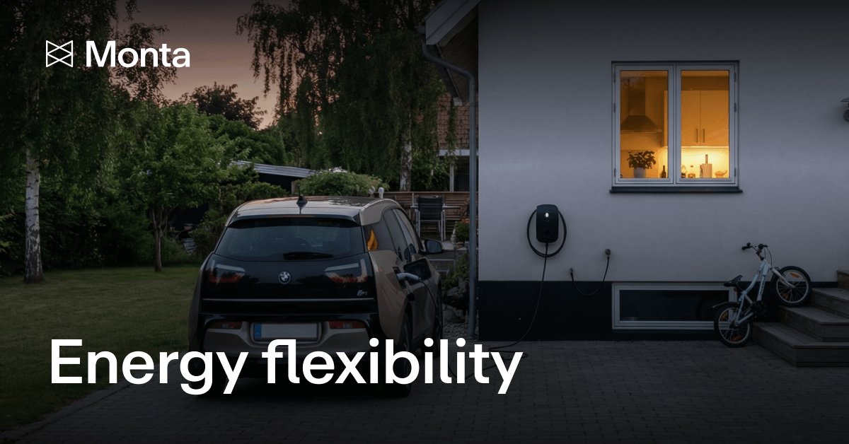 A dark-coloured electric vehicle parked in a driveway beside a house, with an EV charger on the wall and warm light glowing from the window.