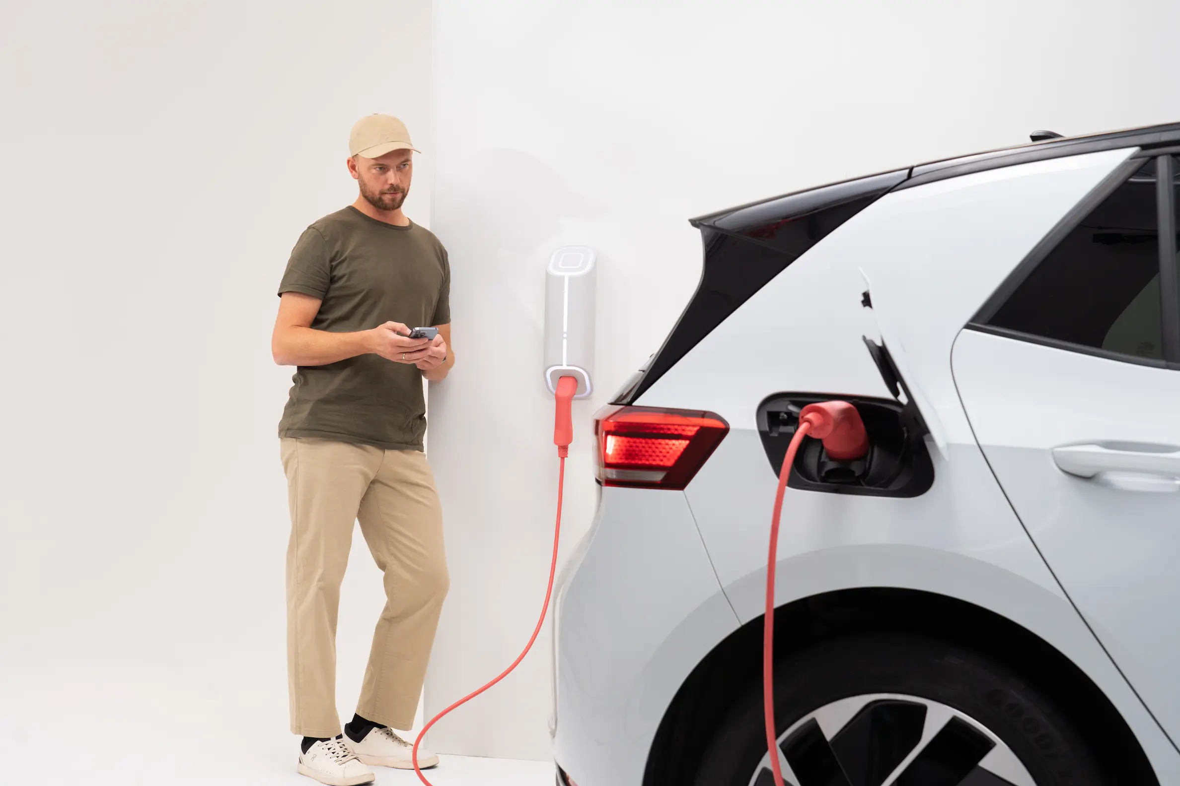 A man in a cap checks his phone beside a white electric hatchback plugged into a wall-mounted home charger by a red cable.