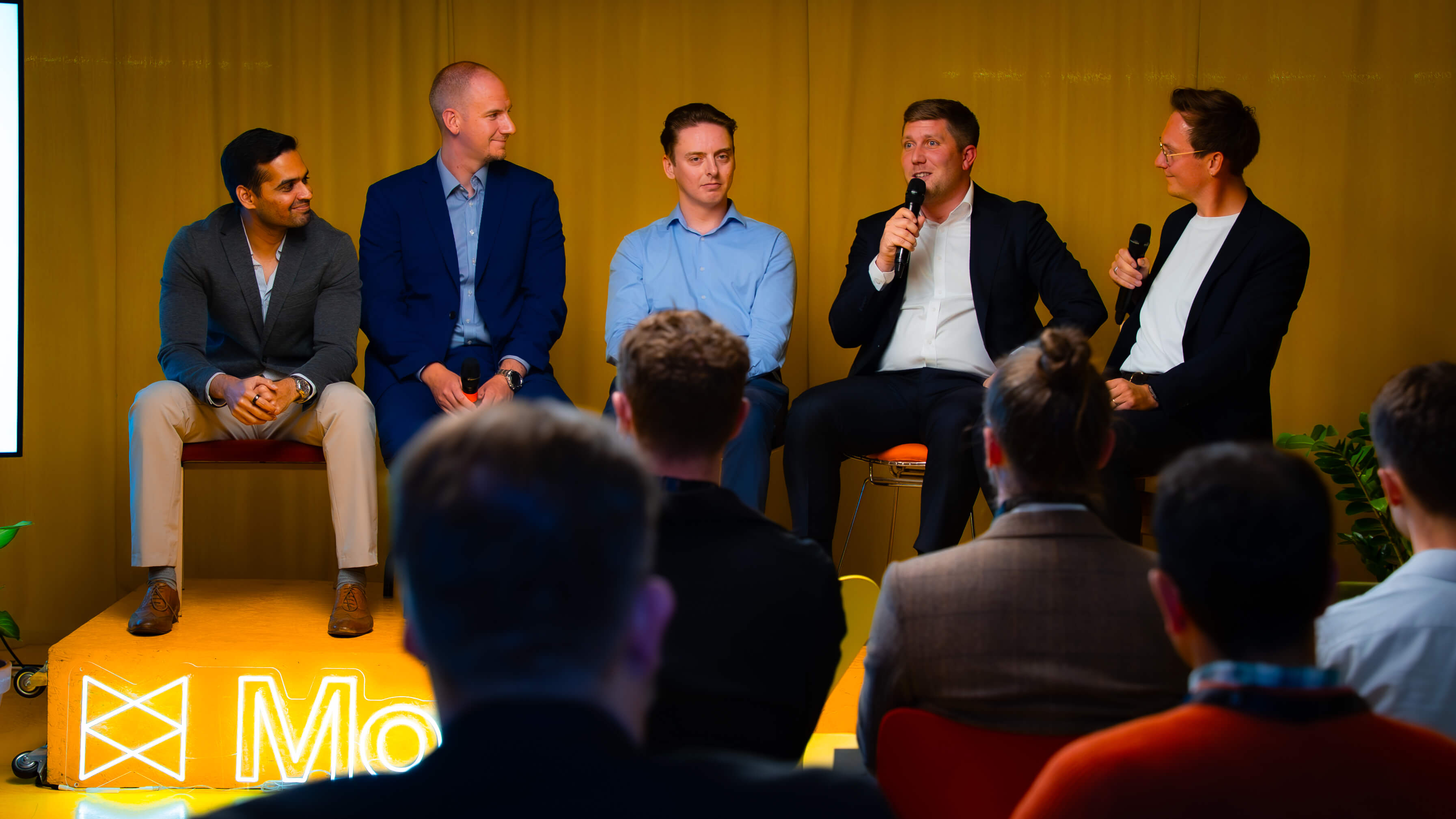 A panel discussion featuring four speakers in suits, seated on chairs against a yellow backdrop, with an audience visible in the foreground.
