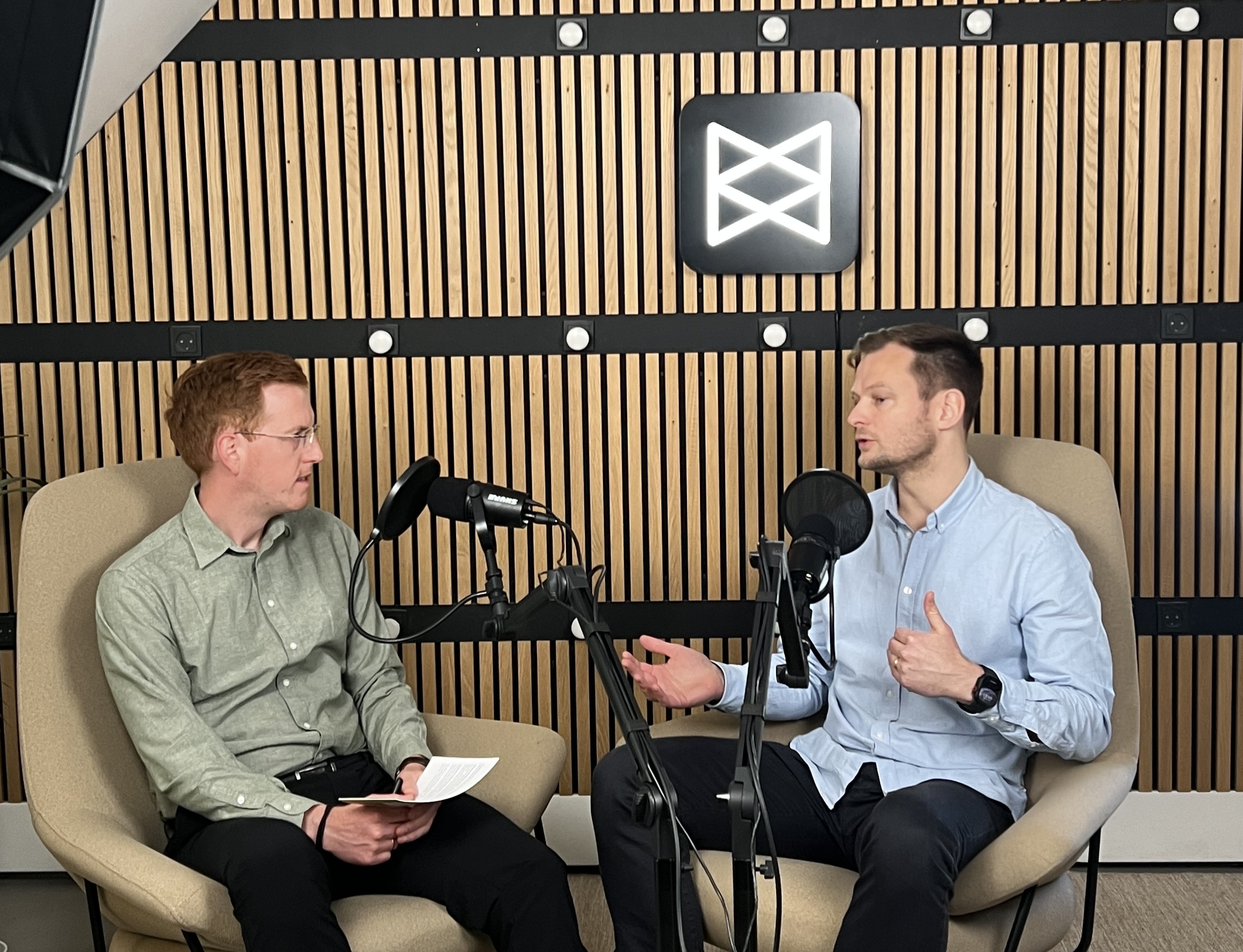 Two men sit in armchairs talking into podcast microphones in front of a wooden slat wall with an illuminated Monta logo.