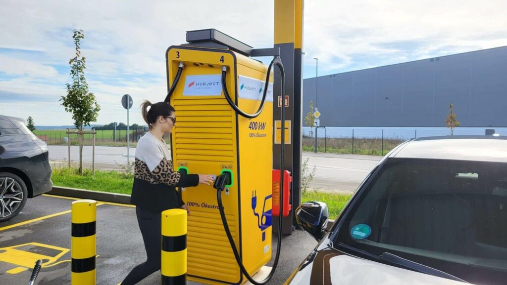 A woman handling the charging cable at a yellow 400 kW Hubject-branded DC fast charger, with the BMW test vehicle parked alongside