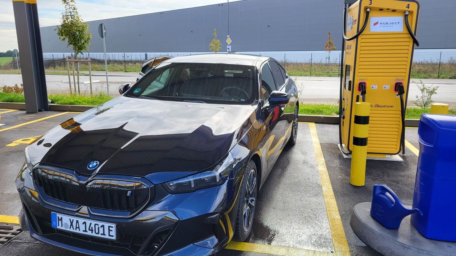 A black BMW electric saloon parked beside a yellow 400 kW DC fast charger carrying Hubject Plug&Charge branding.