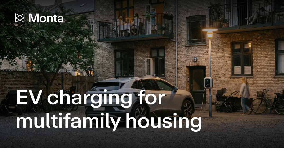 A modern electric vehicle parked near a charging station outside a multifamily housing complex, with warm lighting and greenery.