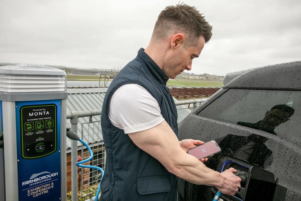 A man holds a phone while plugging a charging cable into a black car at a Monta-powered charge point at Farnborough International