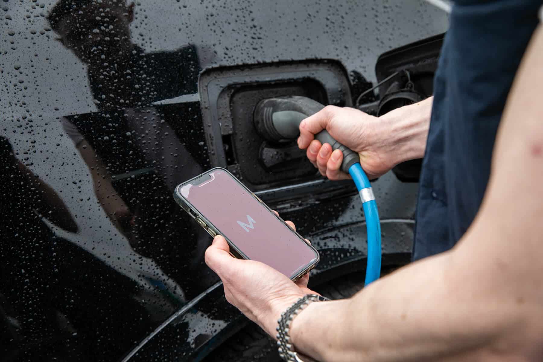 Close-up of a person holding a phone running the Monta app while plugging a blue charging cable into a rain-covered black car.