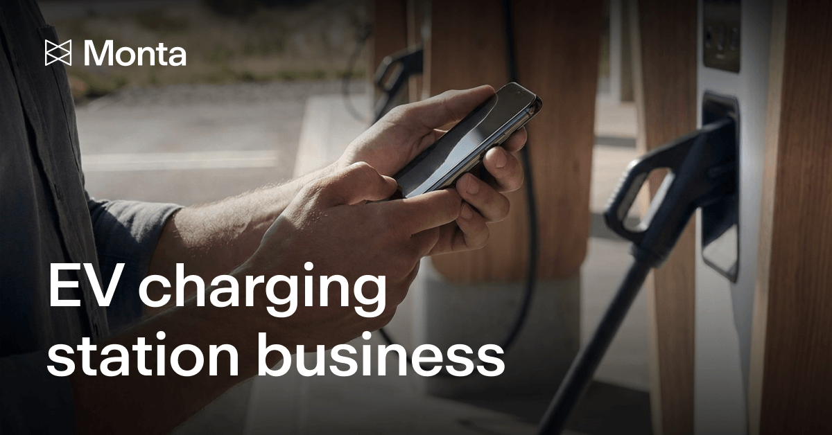 A person uses a smartphone while standing near an EV charging station, with charging cables visible in the background.