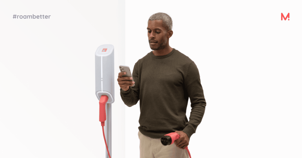 A man holding a phone in one hand and a red charging connector in the other, standing beside a white wall-mounted charge point.