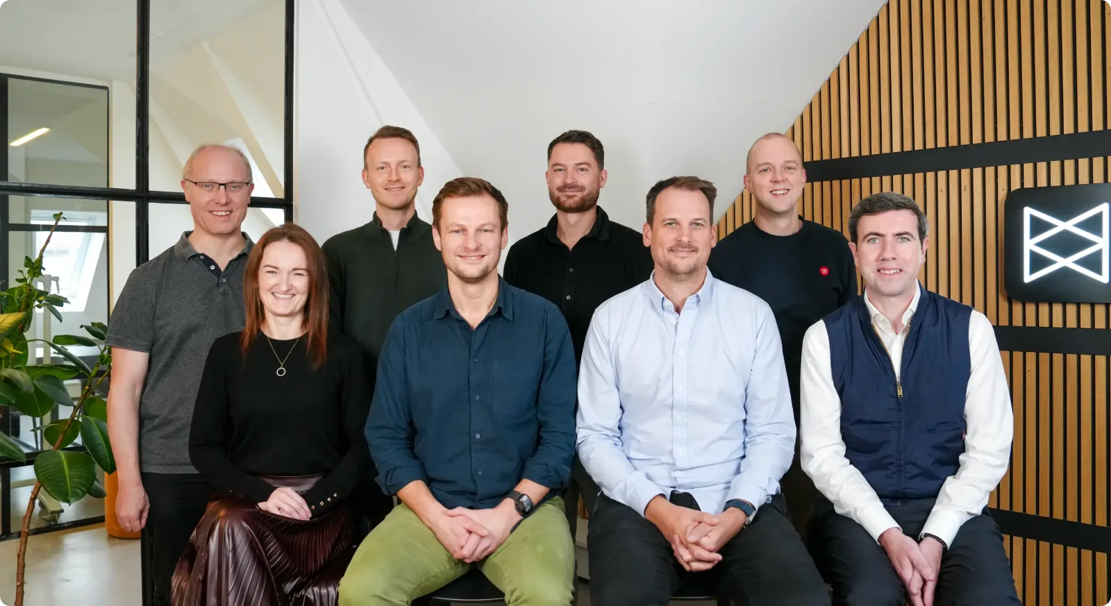 Monta’s leadership team, eight people posed in the Copenhagen office in front of a wooden wall with the Monta logo.